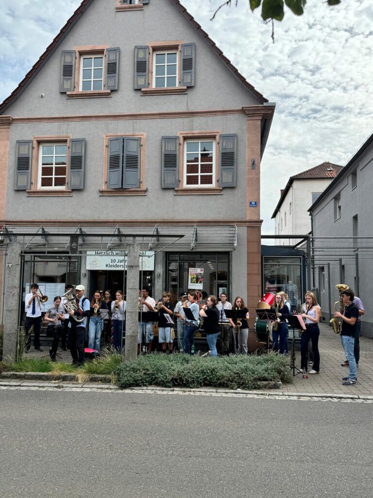 Die United Concert Band vor einem Haus an der Hauptstraße in Herxheim, im Schaufenster das Plakat des Theaterbummels.